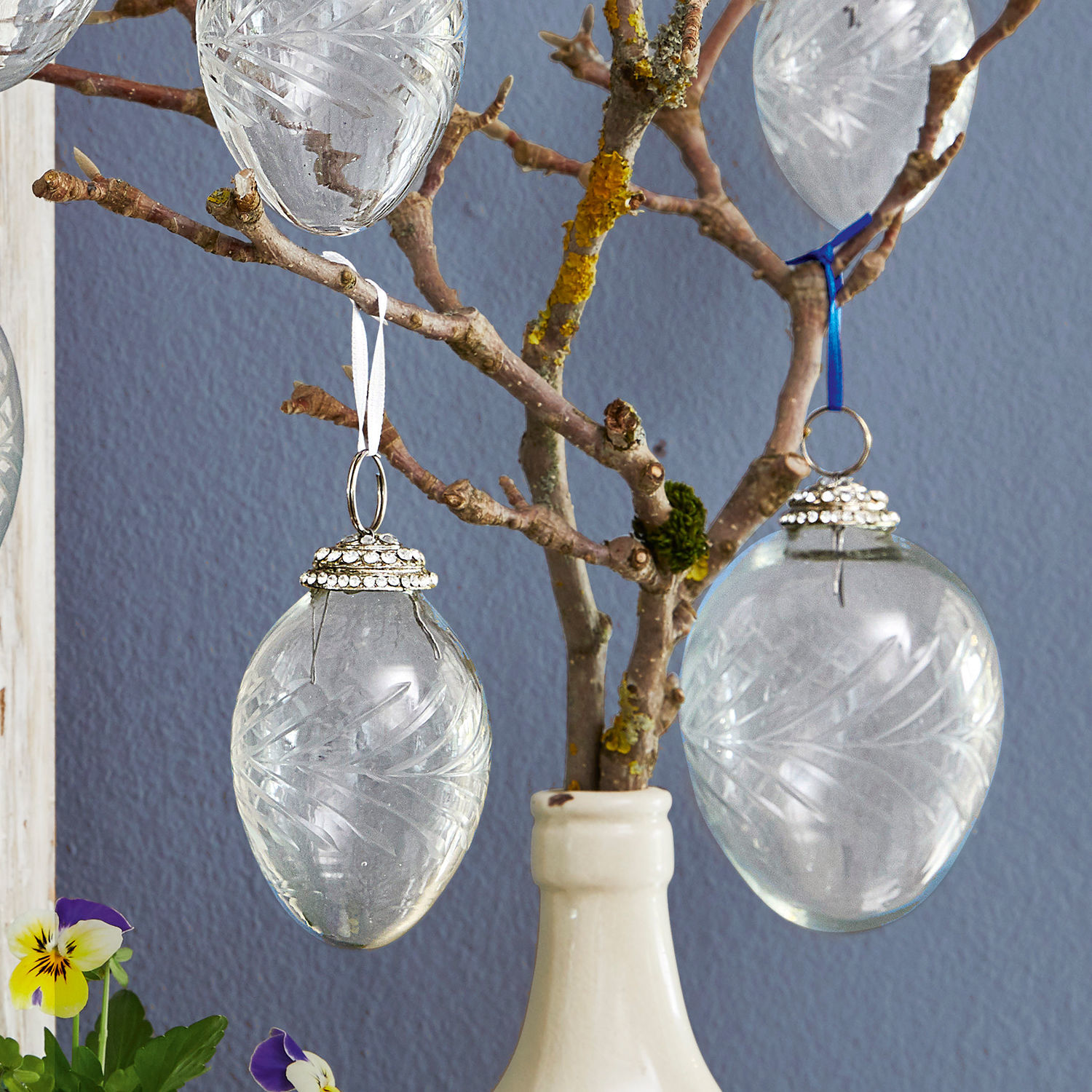 Transparente Glas-Ostereier mit Gravurmuster an Zweig in weißer Keramikvase hängend