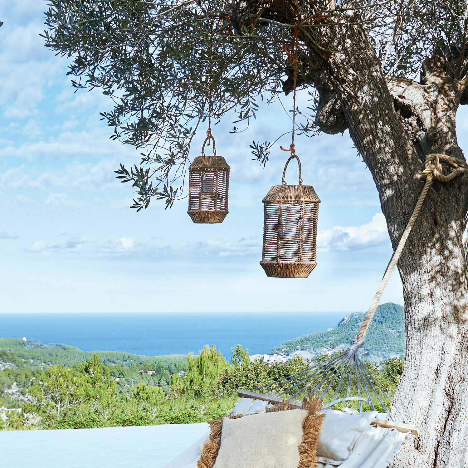 Zwei geflochtene Laternen hängen an einem Baum über einer Hängematte mit Kissen, Meer- und Waldblick