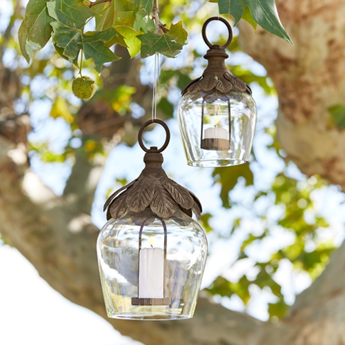 Glaslaternen mit Metallverzierung und weißen Kerzen hängen an einem Baum als Gartendekoration