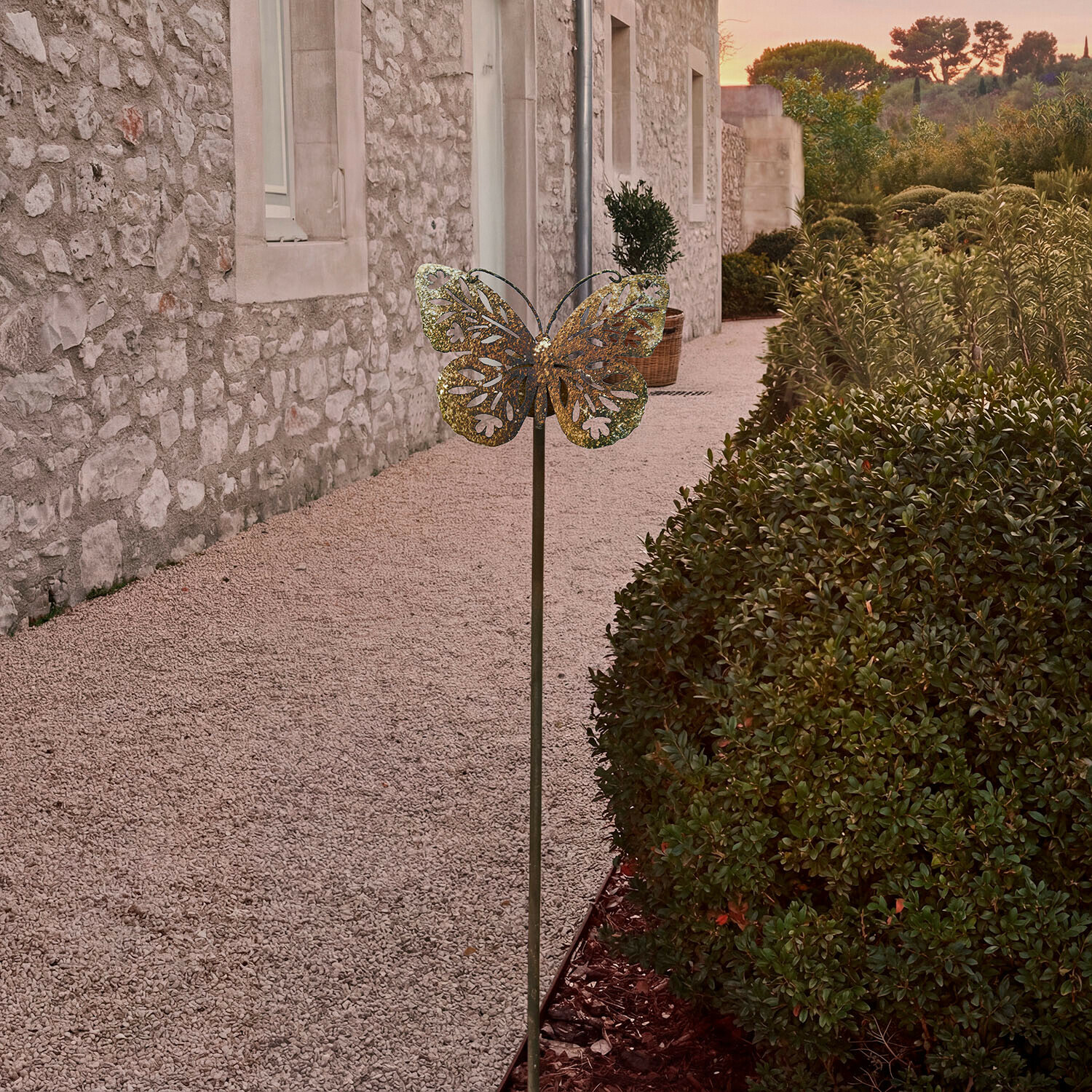 Metall-Gartendekoration in Schmetterlingsform in Messingfarbe vor Steinmauer und grünen Büschen