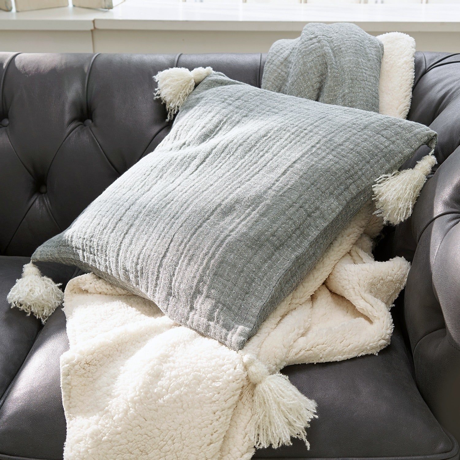 Graues Kissen mit Quasten und cremefarbene weiche Decke auf dunklem Ledersofa im Wohnzimmer