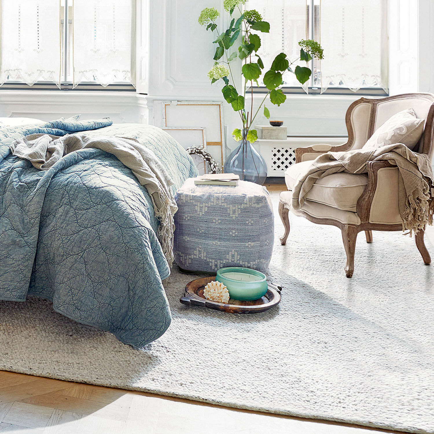 Gemütliches Schlafzimmer mit blauem Bettüberwurf, beigem Sessel und weichem Teppich auf Holzboden