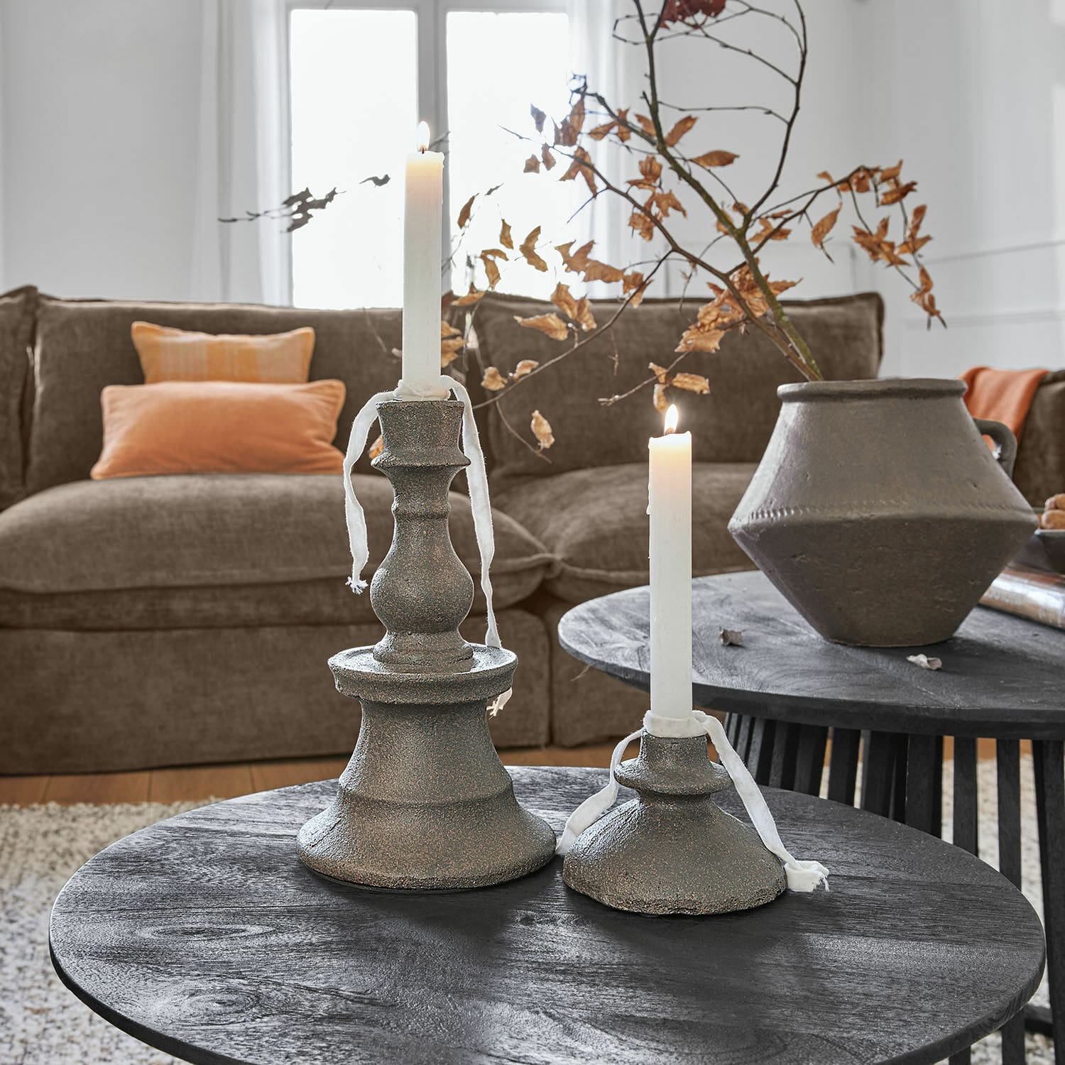 Zwei graue Kerzenhalter mit weißen Kerzen auf schwarzem Holztisch im Wohnzimmer mit braunem Sofa