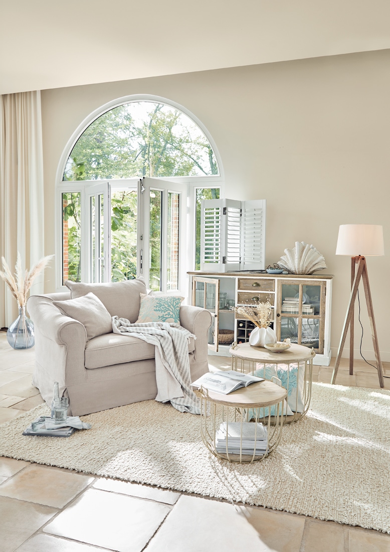 Gemütlicher beiger Sessel mit Kissen und Decke in hellem Wohnzimmer mit Fenster und Stehlampe