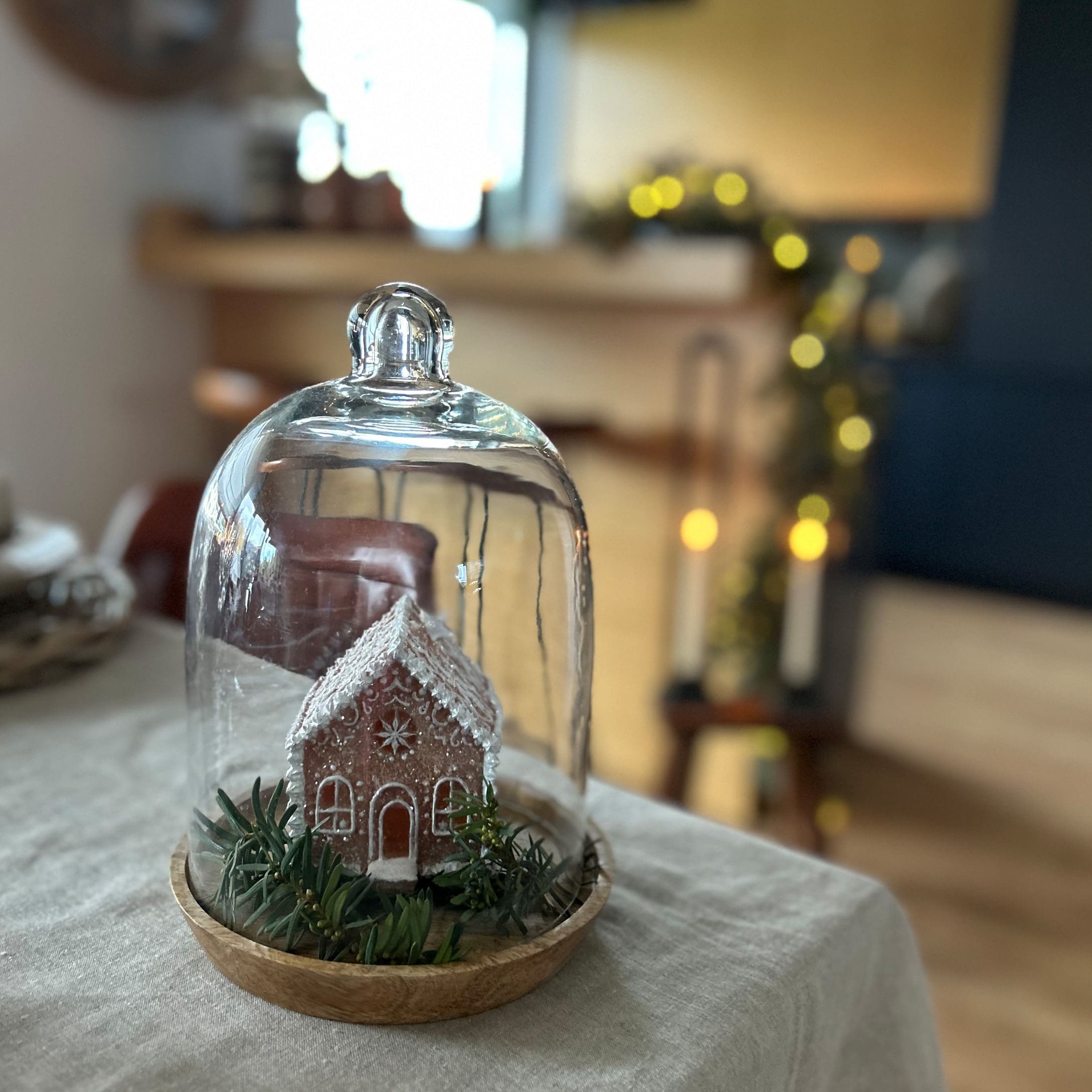 Dekoratives Lebkuchenhaus unter Glaskuppel auf Holzplatte mit grünen Zweigen als Deko