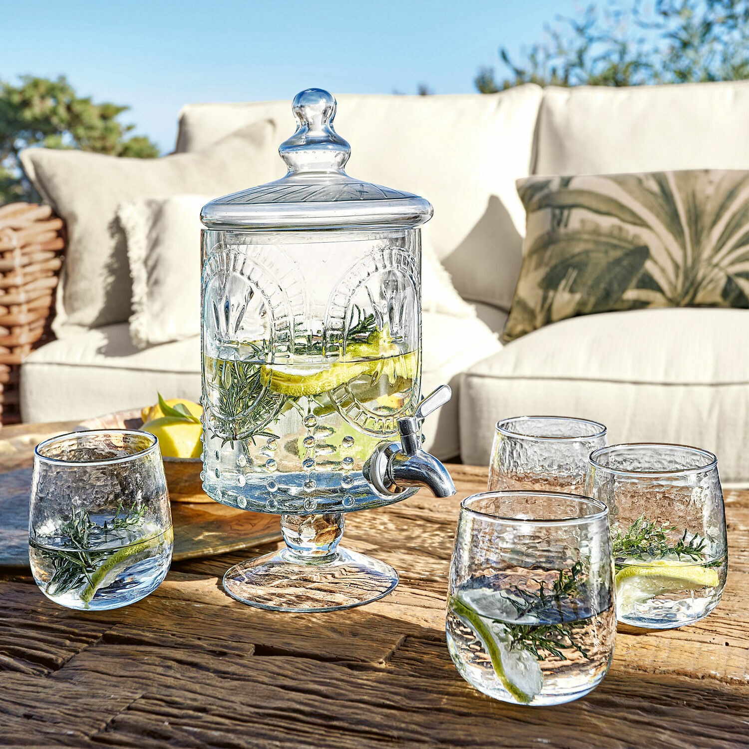 Glas-Getränkespender mit Deckel und Zapfhahn auf Holztisch mit vier Gläsern Wasser mit Zitrone