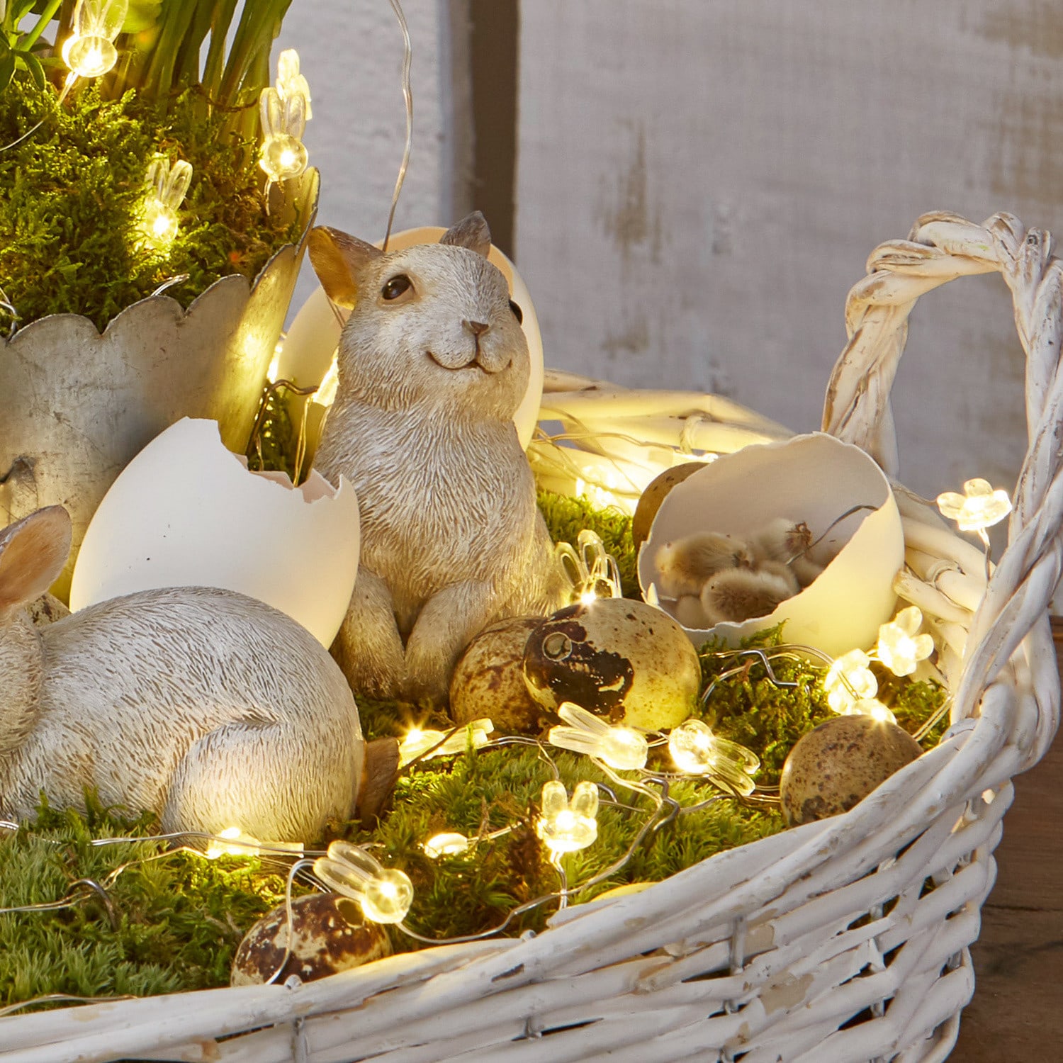 Weißer Korb aus Weide mit Hasenfiguren, Eiern und LED-Lichterkette auf grünem Moos dekoriert