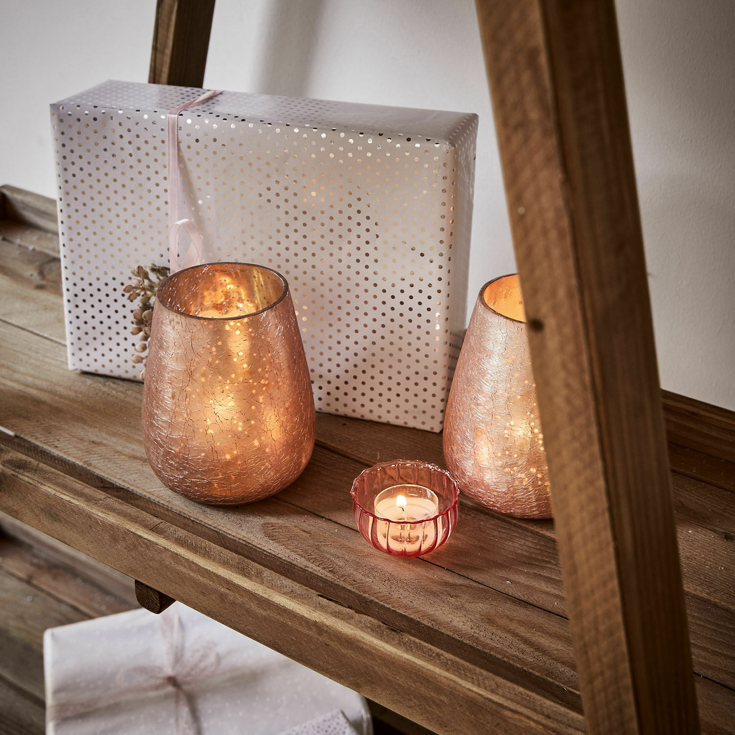 Glas-Teelichthalter in Roségold auf Holzregal mit Geschenk im Hintergrund