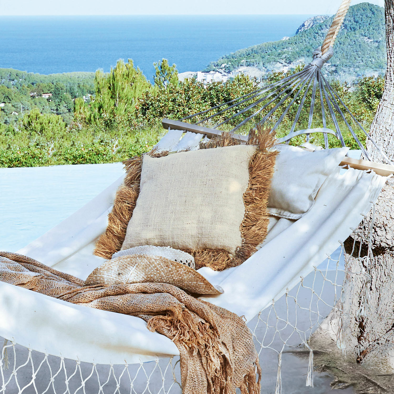 Weißes Hängemattensofa mit natürlichem Kissen und beiger Decke vor grünem Meerblick