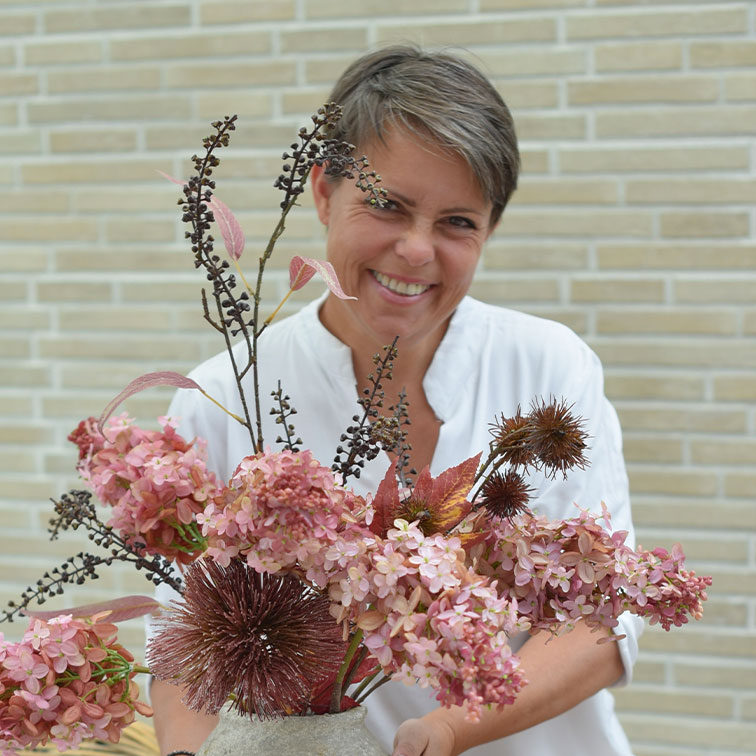 Frau in weißer Bluse hält Vase mit rosa und braunen getrockneten Dekoblumen