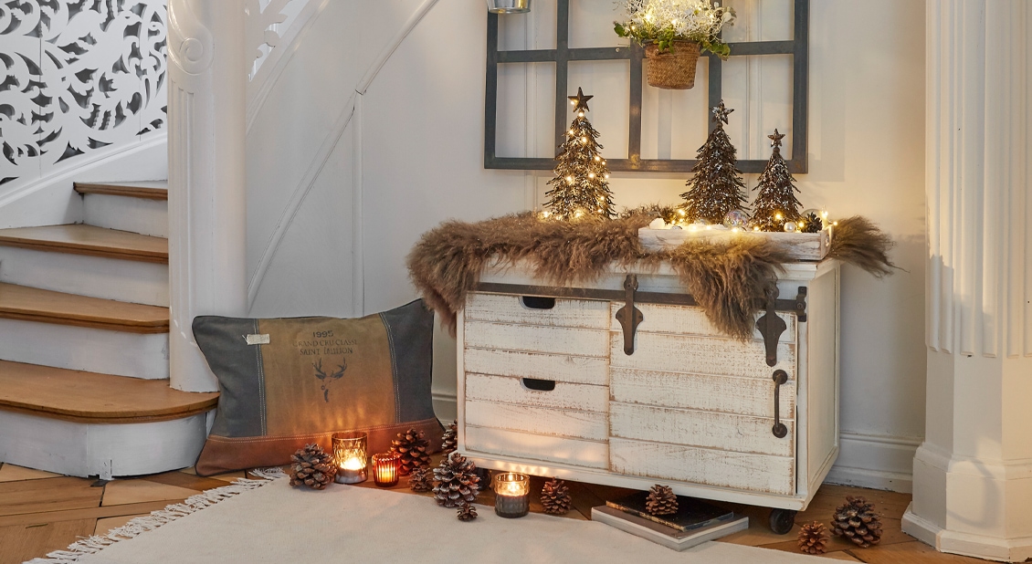 Gemütliche Winterdekoration mit beleuchteten Tannenbäumen und Naturzapfen auf weißem Holzkommode