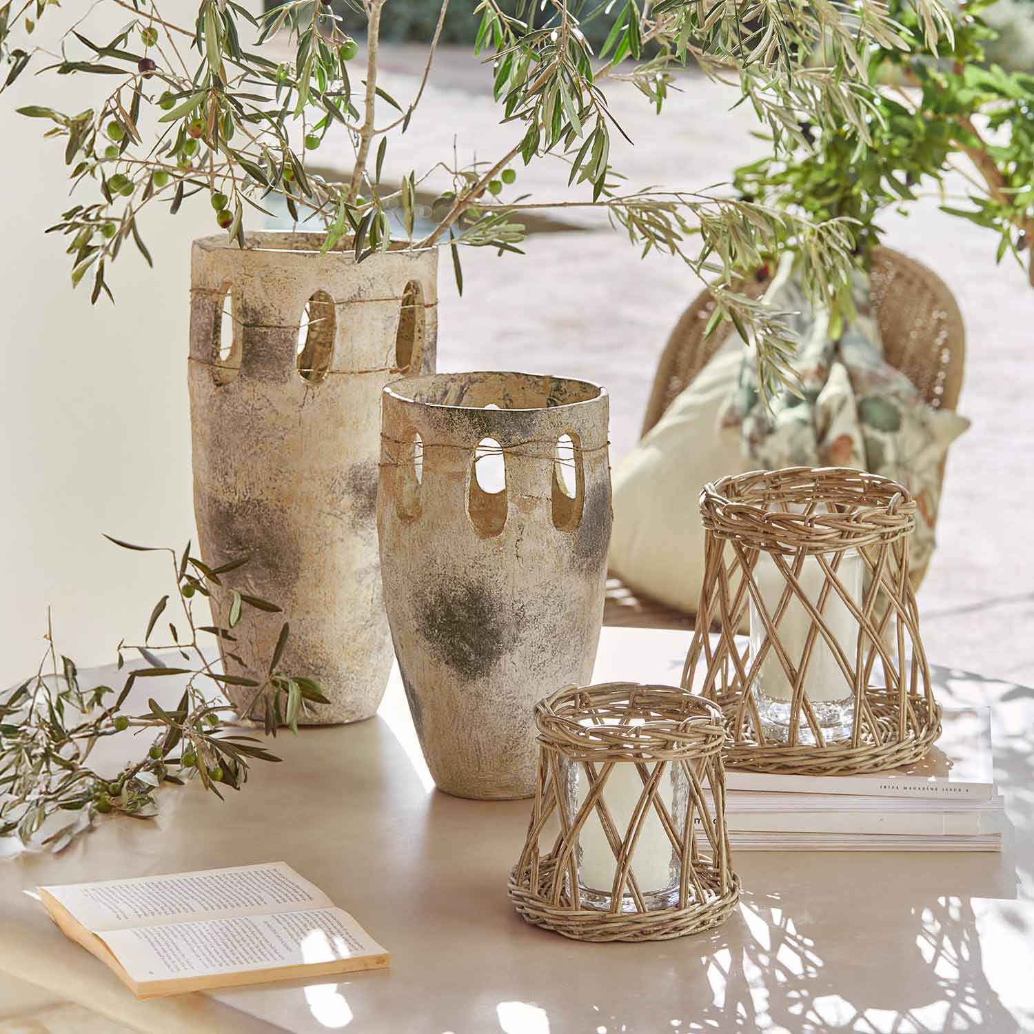 Zwei große Keramikvasen und zwei Rattan-Laternen auf einem Tisch mit grünen Zweigen und einem offenen Buch