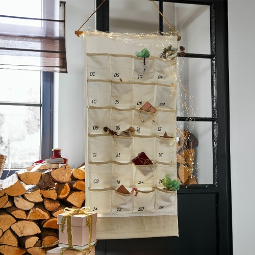 Beiger Adventskalender aus Stoff mit nummerierten Taschen und Dekorationen, hängend an schwarzer Tür in gemütlichem Raum