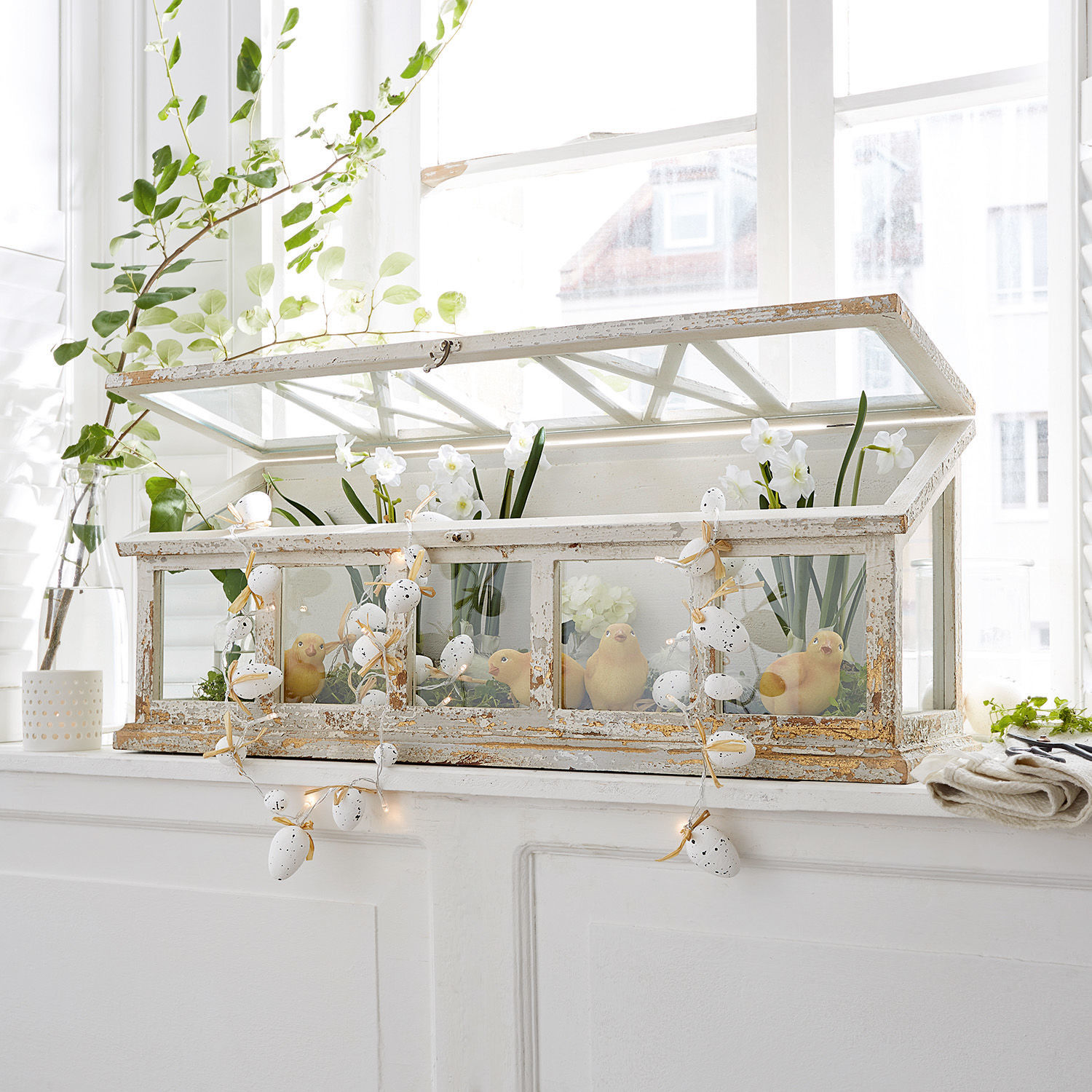 Weiße Vintage-Holzvitrine mit Glas und Frühlingsdeko, LED-Lichterkette auf Fensterbank