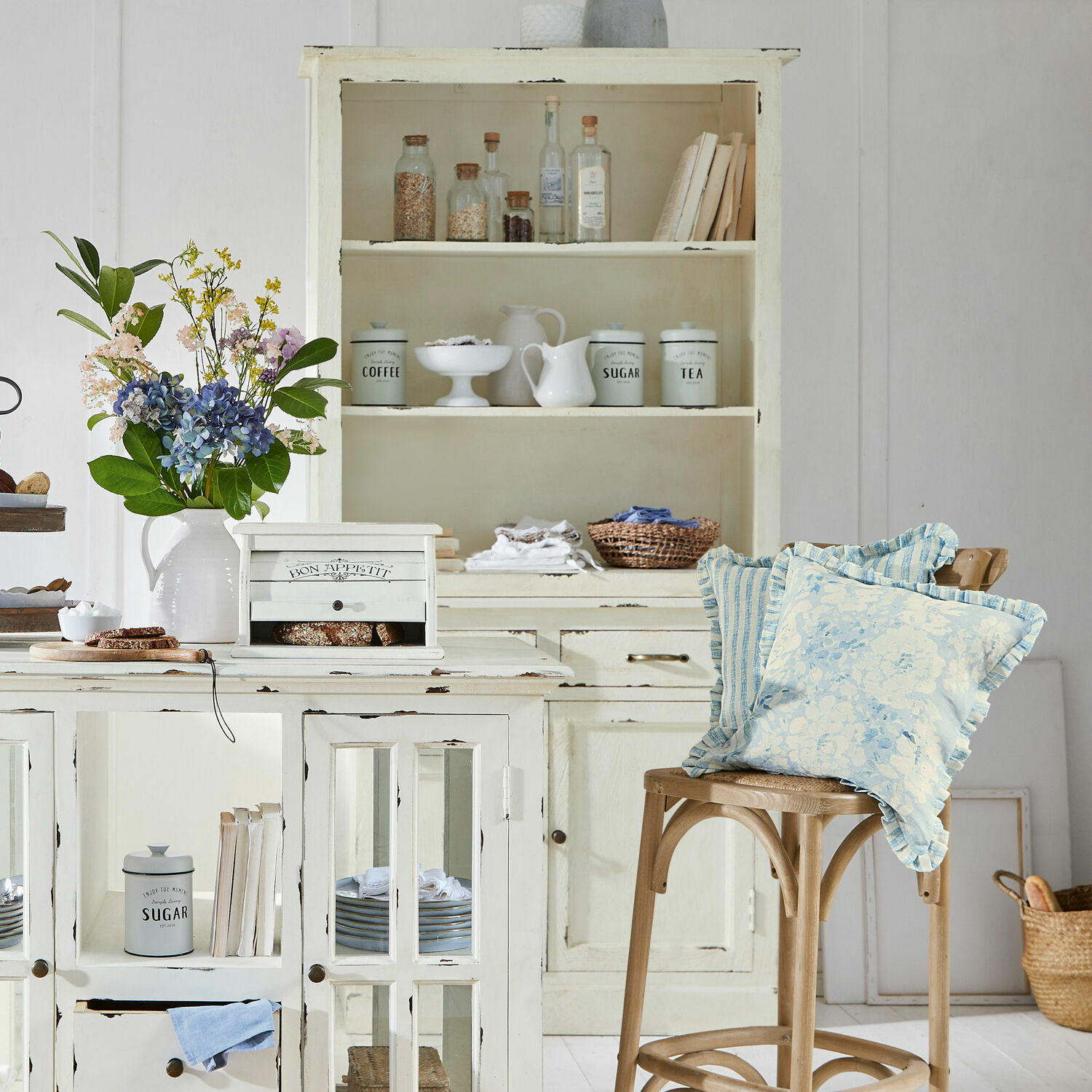 Kremfarbener Holz-Küchenschrank mit Shabby-Chic-Effekt und blauen dekorativen Kissen auf Holzstuhl