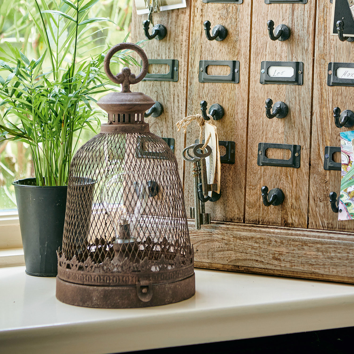 Rustikale Metalllampe mit Rostpatina und Gitter auf weißem Fensterbrett vor Holzschrank und Pflanze