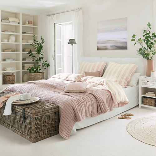 Gemütliches Schlafzimmer mit weißer Bettwäsche und rosa Decke, natürliche Dekorationen und Korbtruhe