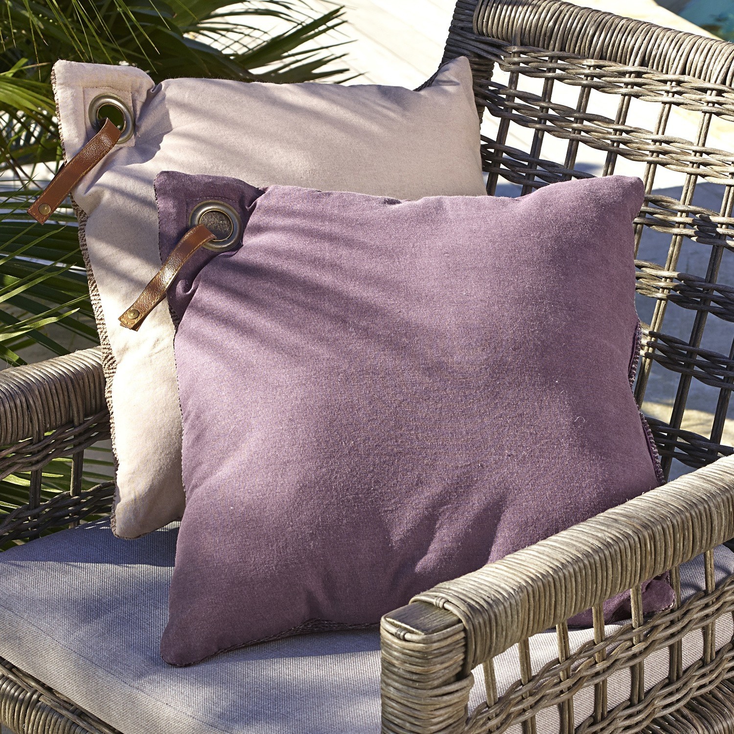 Zwei dekorative Kissen aus weichem Stoff in Beige und Violett auf einem Korbsessel im Außenbereich