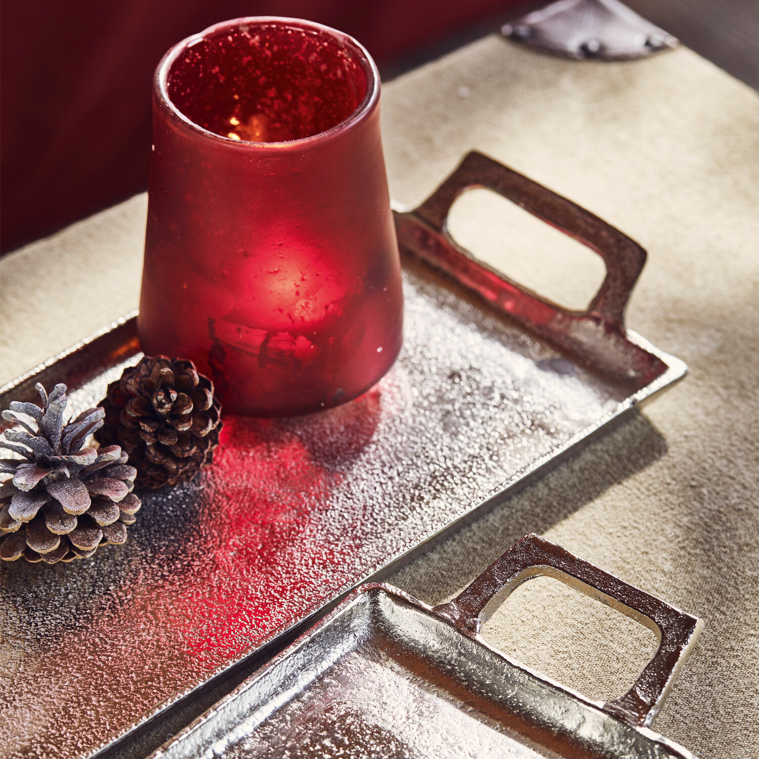 Roter Glas-Teelichthalter auf Metalltablett mit dekorativen Zapfen auf beigem Hintergrund