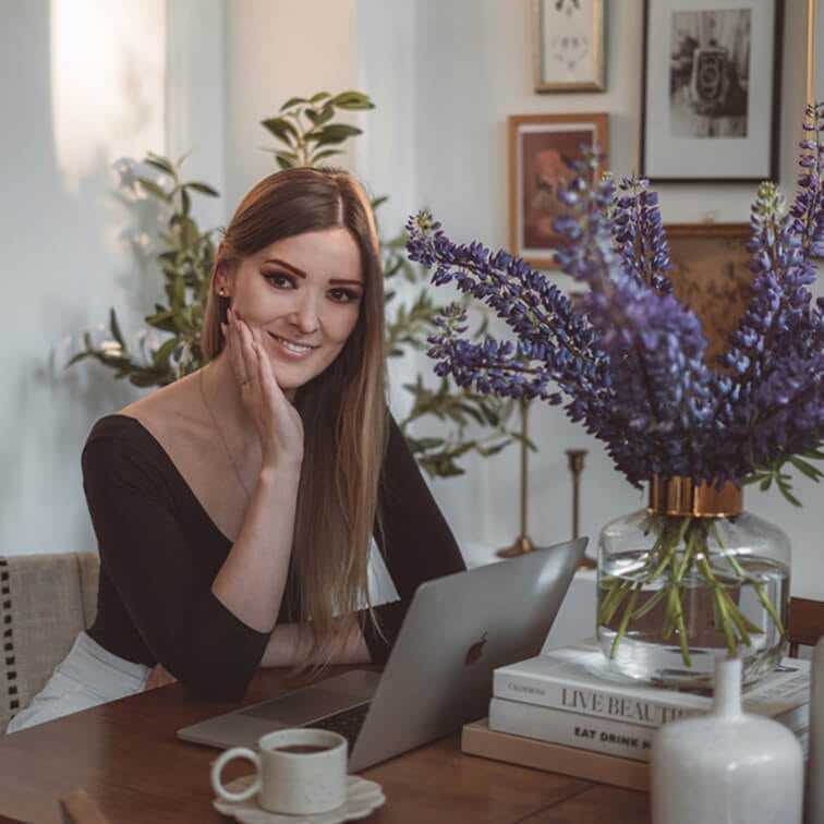 Frau arbeitet am Laptop an Holztisch mit lila Blumenstrauß und Kaffeetasse