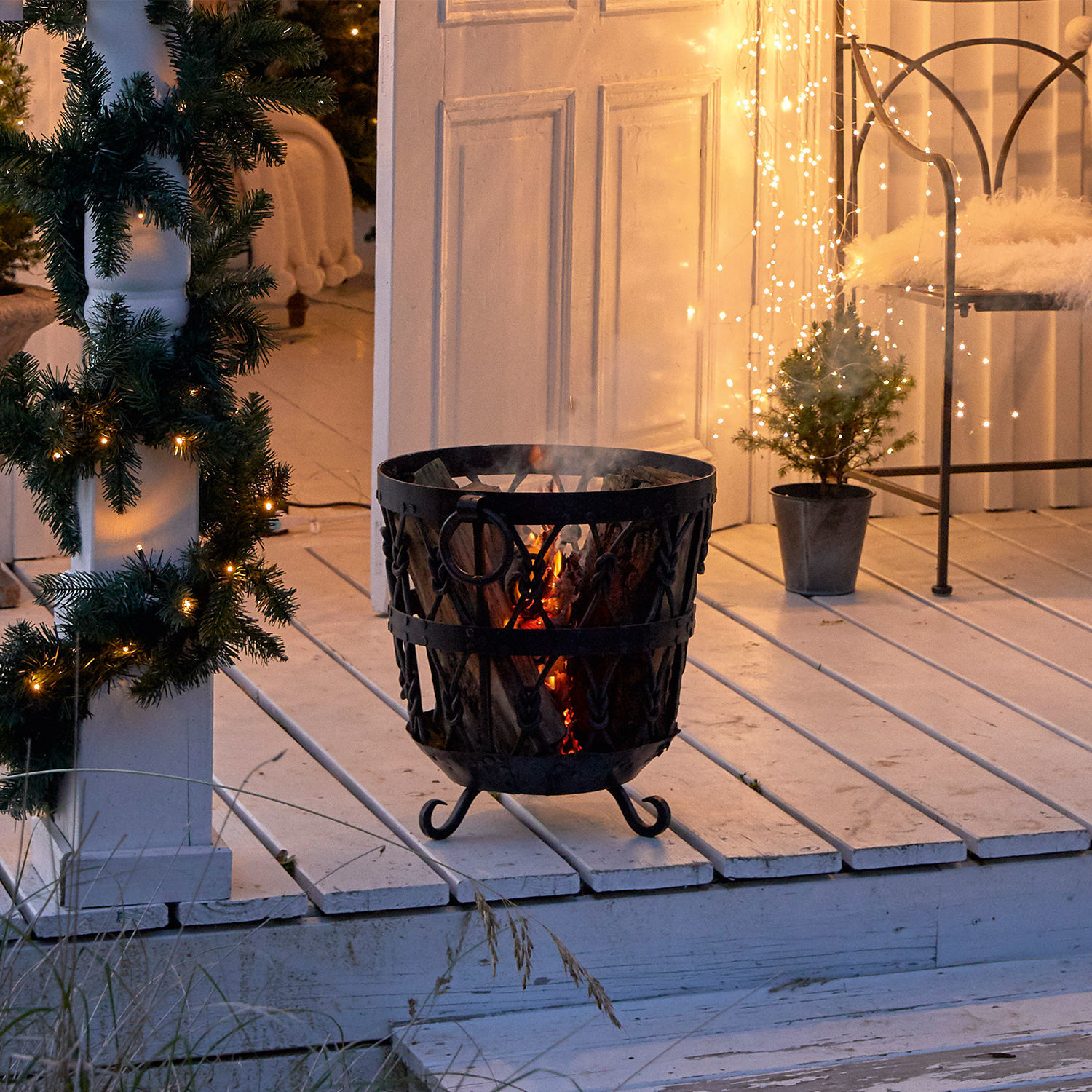 Schwarzer Metall-Feuerkorb mit brennendem Holz auf weißem Holzdeck mit Weihnachtsdekoration