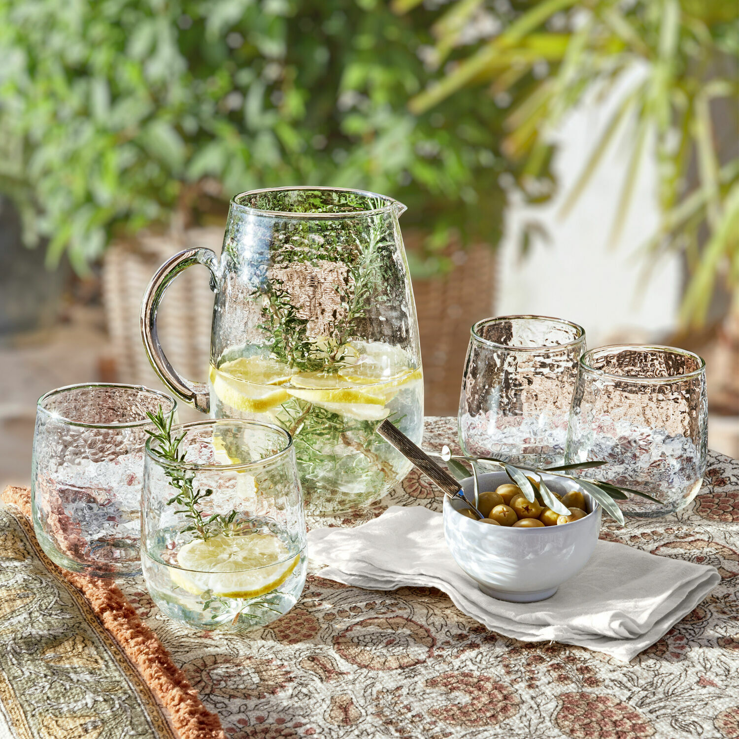 Glaskrug und vier strukturierte Gläser mit Zitronenwasser und Kräutern auf gemustertem Tischtuch