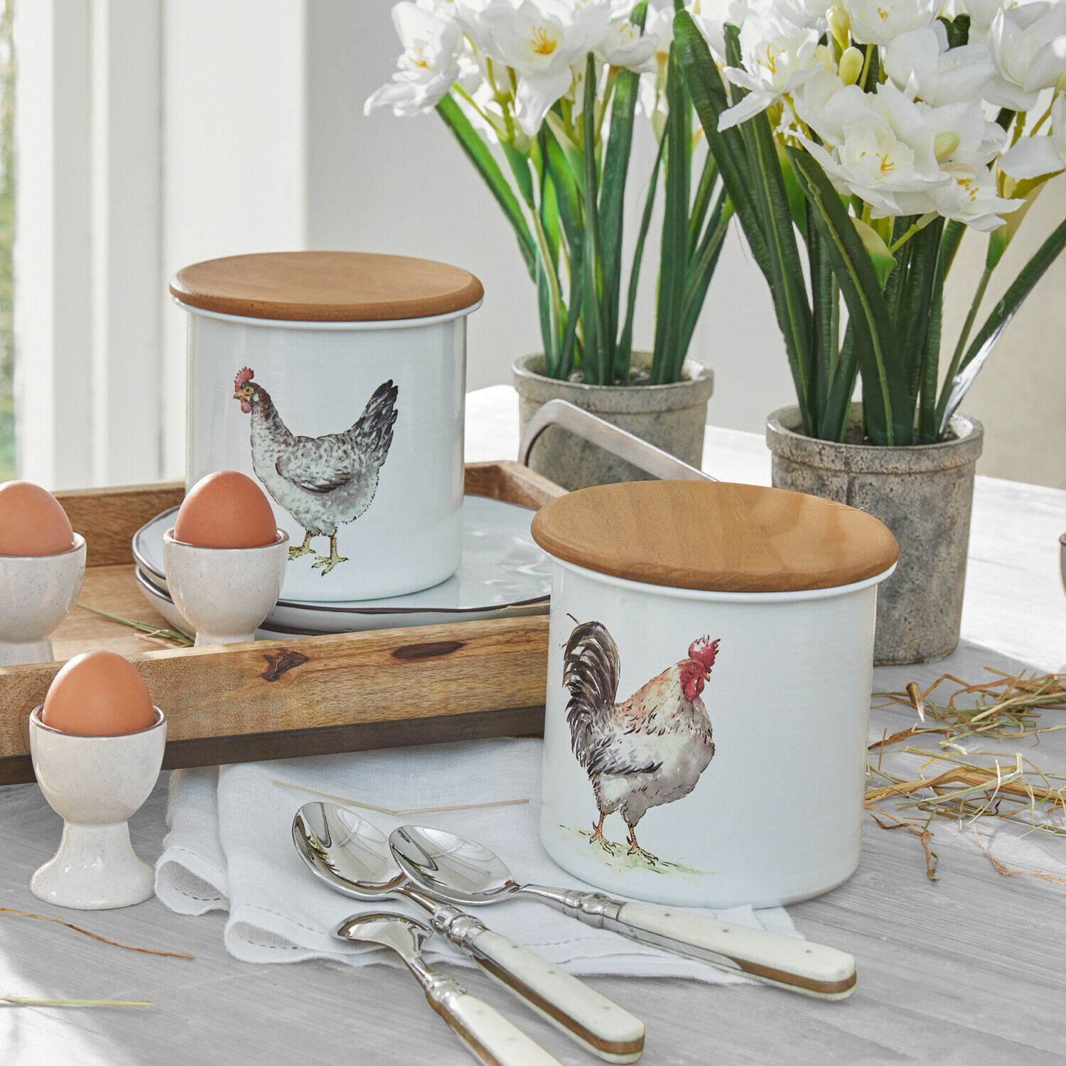 Weiße Keramikbehälter mit Holzdeckeln und Huhn- sowie Hahn-Motiv auf Küchentisch mit Besteck und Eiern