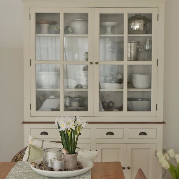Cremefarbener Holzvitrinenschrank mit Glastüren und Tischdeko mit Blumen und Kerzen