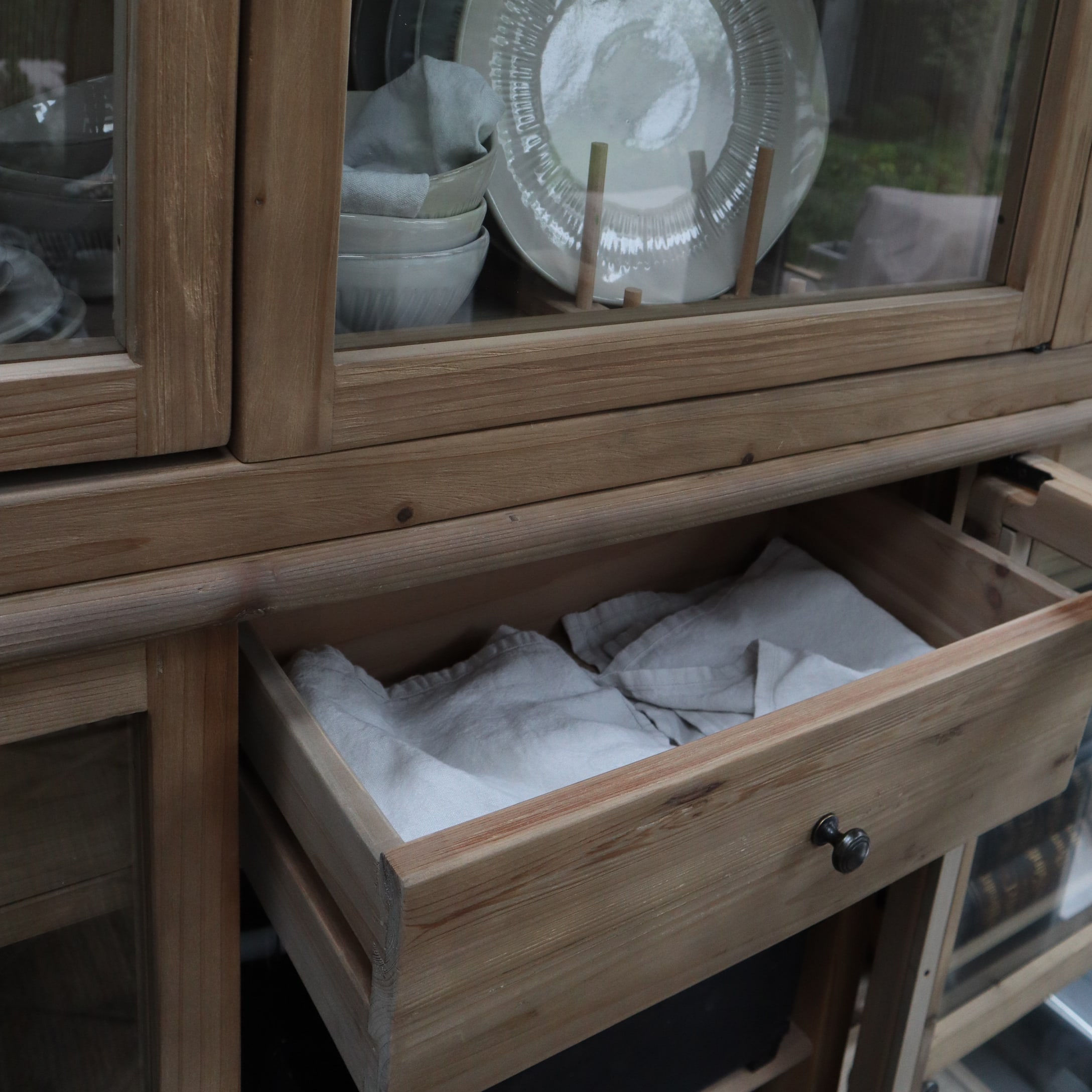 Offene Schublade aus hellem Holz mit Leinenstoff in Vitrine mit Tellern und Schalen