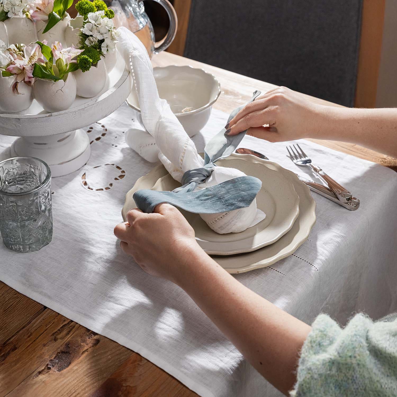 Hände arrangieren weißen Leinentischläufer und Serviette mit blauem Band auf Holztisch mit Porzellantellern