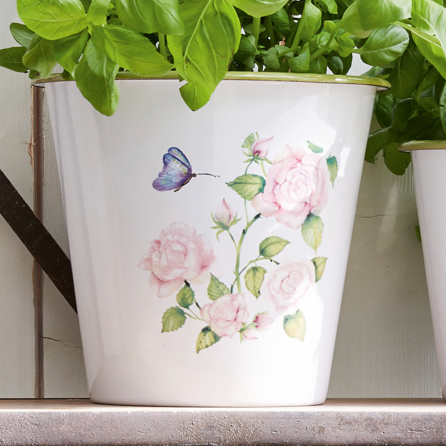 Weißer Metall-Blumentopf mit rosa Blumenmotiv und grünen Pflanzen darin