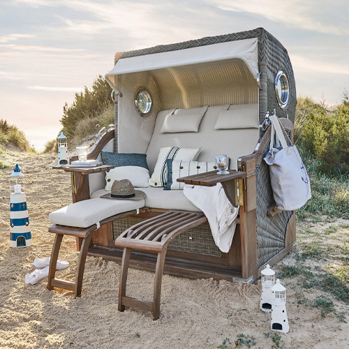 Strandkorb aus Rattan mit beigen Polstern und Holzfußstützen im Sand, ideal zum Entspannen