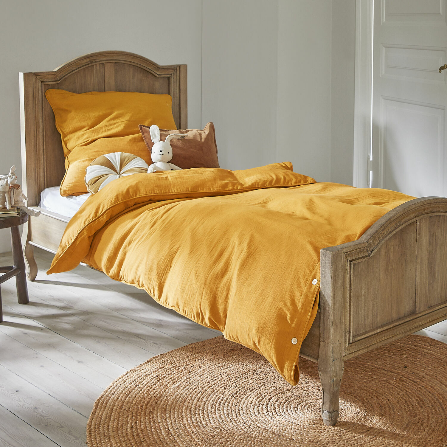 Holz-Bett mit senfgelber Bettwäsche, weichen Kissen und Hasenplüschtier in hellem Raum