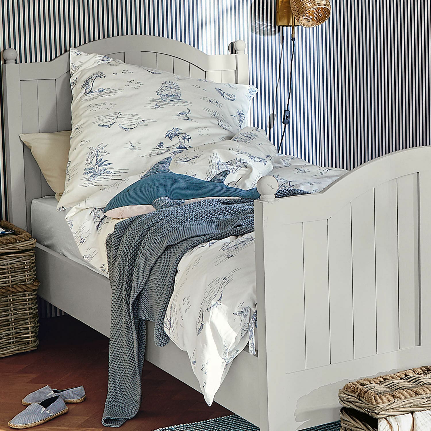 Weißes Holz-Bett mit maritim gemusterter Bettwäsche und blauem Strickdecke in gemütlichem Schlafzimmer