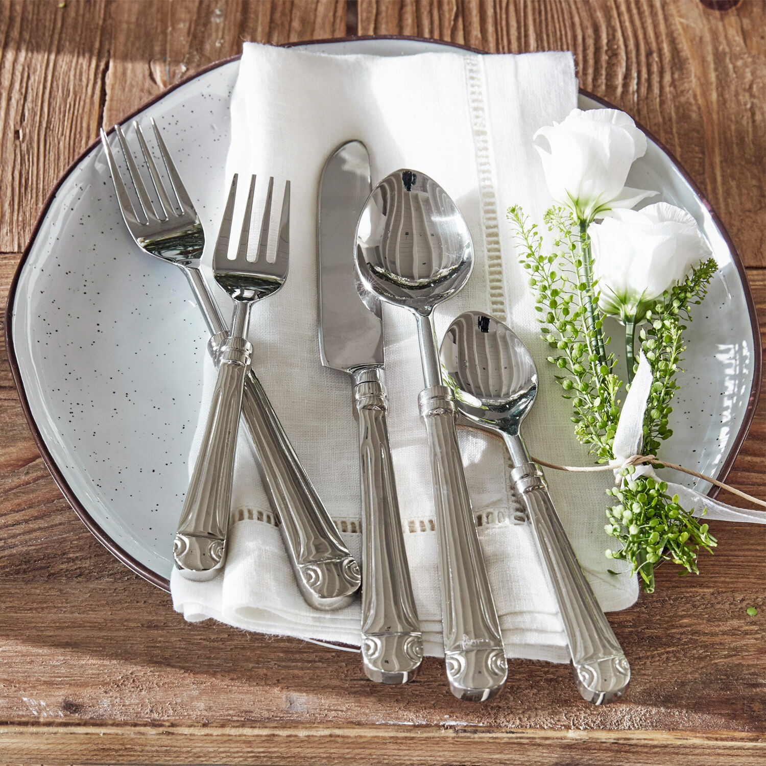 Besteck Eriat Set aus silbernem Besteck auf weißem Stoff mit Blumenarrangement auf Holztisch