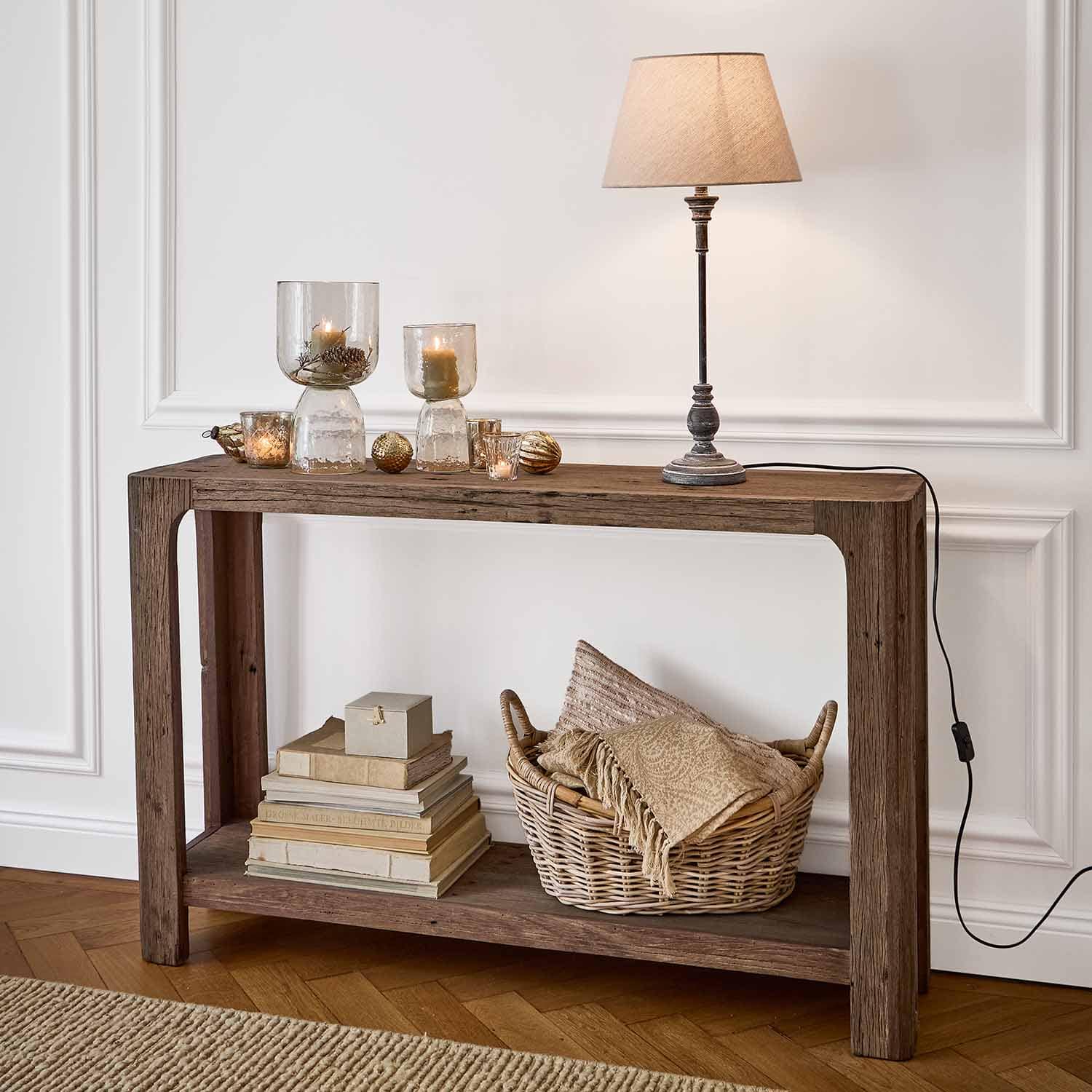 Holzkonsole mit Tischlampe, Kerzen in Glaswindlichtern und Korb mit Textilien aus Rattan