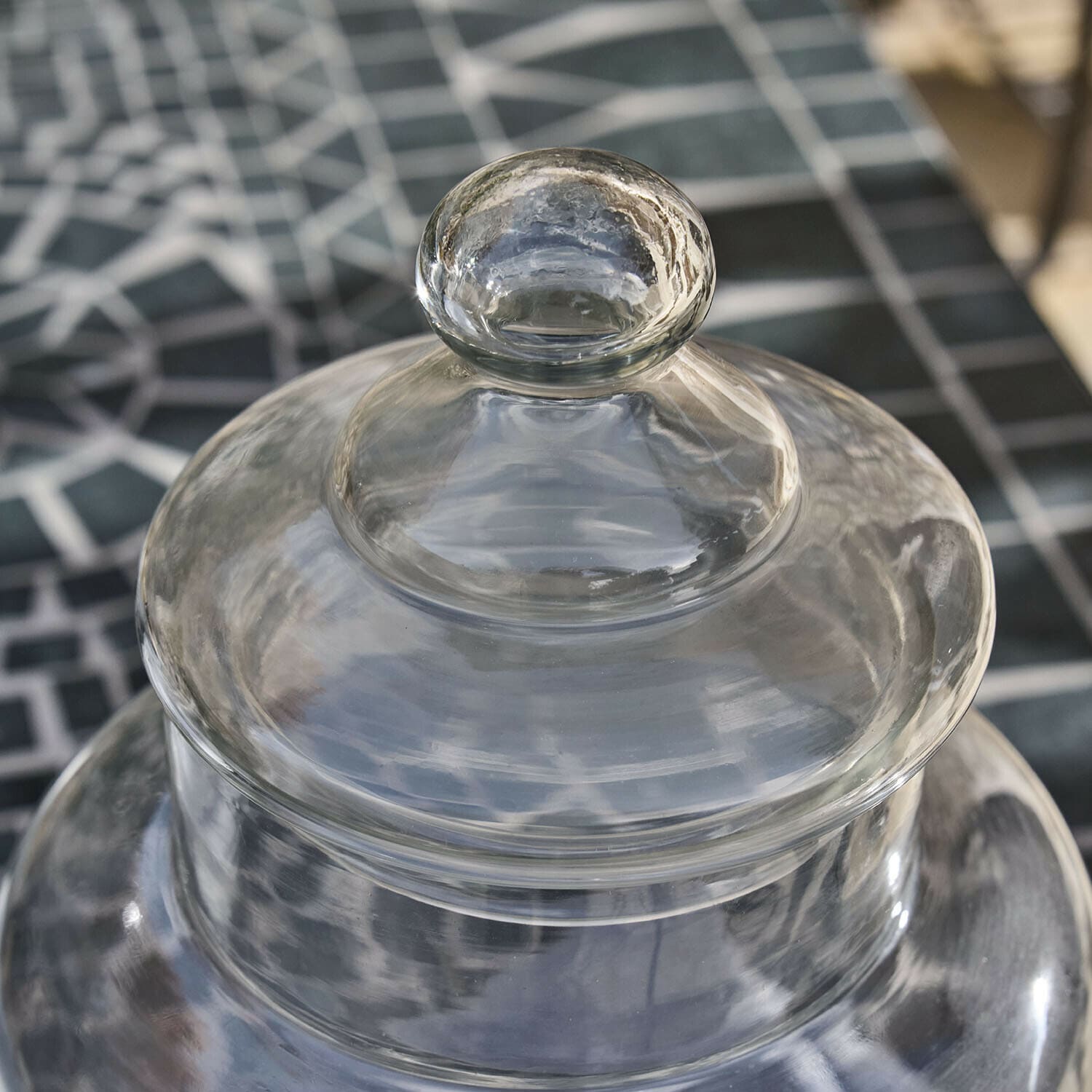 Transparenter Glasdeckel mit rundem Griff vor geometrischem Tischmuster