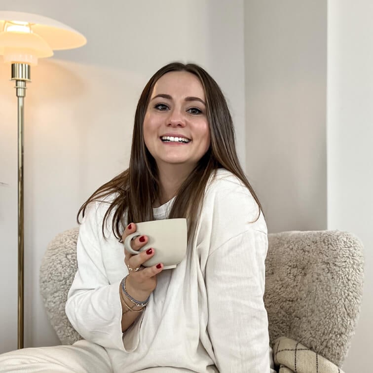Lächelnde Frau im weißen Pullover hält eine beige Tasse und sitzt in einem gemütlichen Sessel in warmem Raum