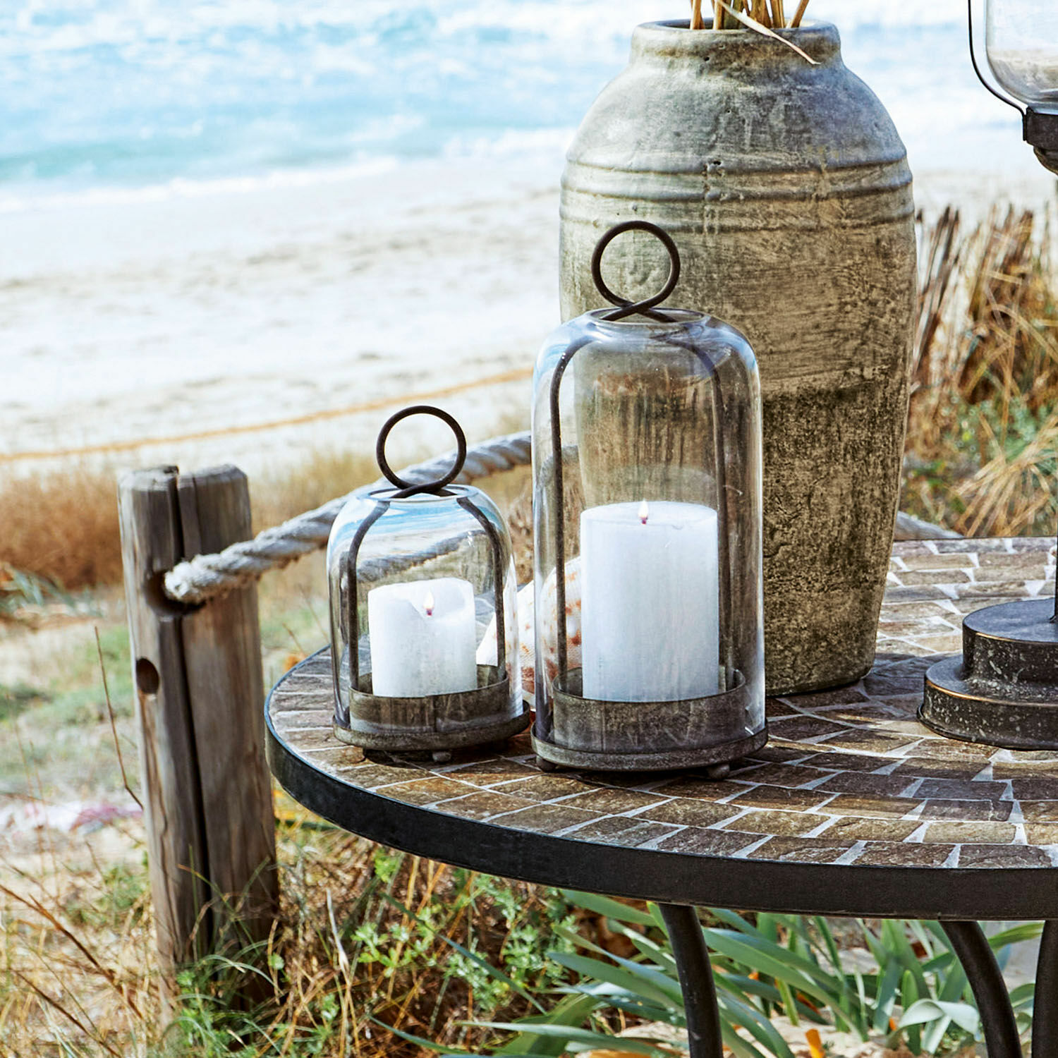 Glaslaternen mit Metallfuß und weißen Kerzen auf rundem Tisch draußen am Meer