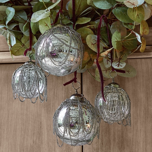 Silberne Glas-Weihnachtskugeln mit dekorativen Ketten vor grünen Blättern und Holzwand hängend