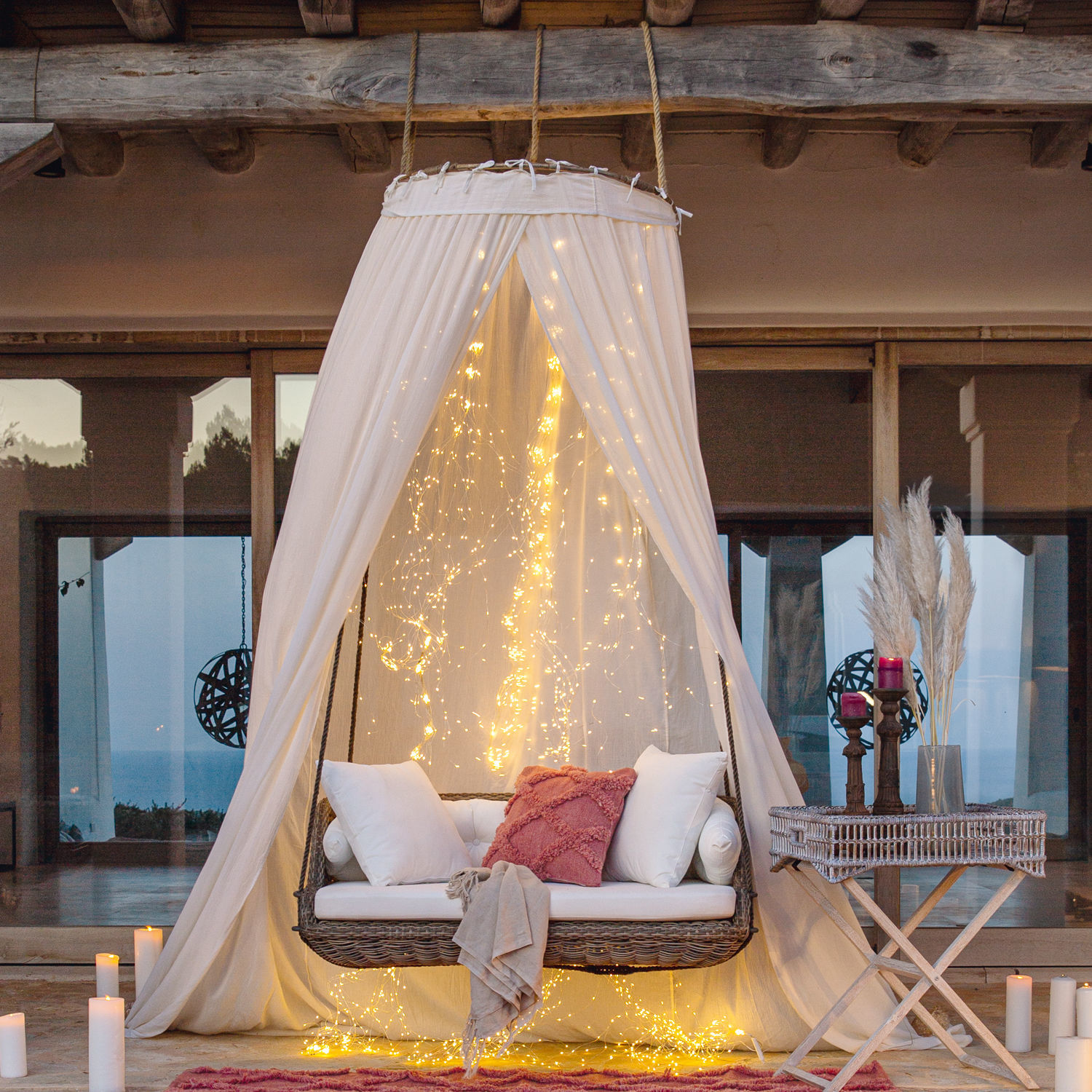 Rattanschaukel mit weißen Kissen und rosa Zierkissen, umgeben von weißem Baldachin und LED-Lichterketten