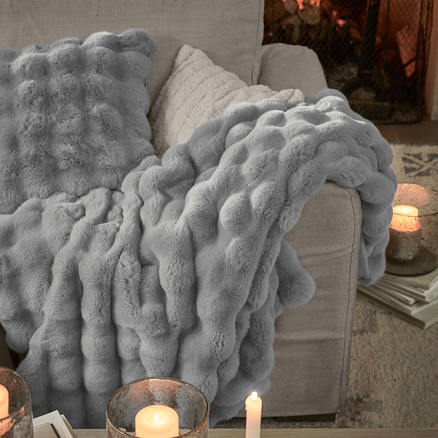 Weiche graue Kunstfell-Decke und Kissen auf beigem Sofa mit Kerzen und Büchern im Hintergrund