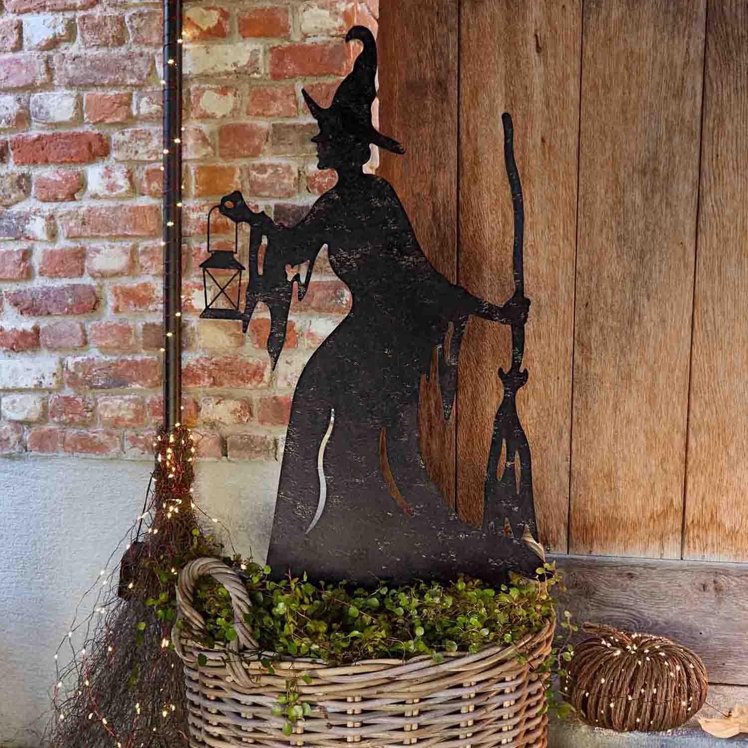 Metall-Dekoration Hexen-Silhouette mit Laterne und Besen im Korb mit Grünpflanzen vor Ziegelwand