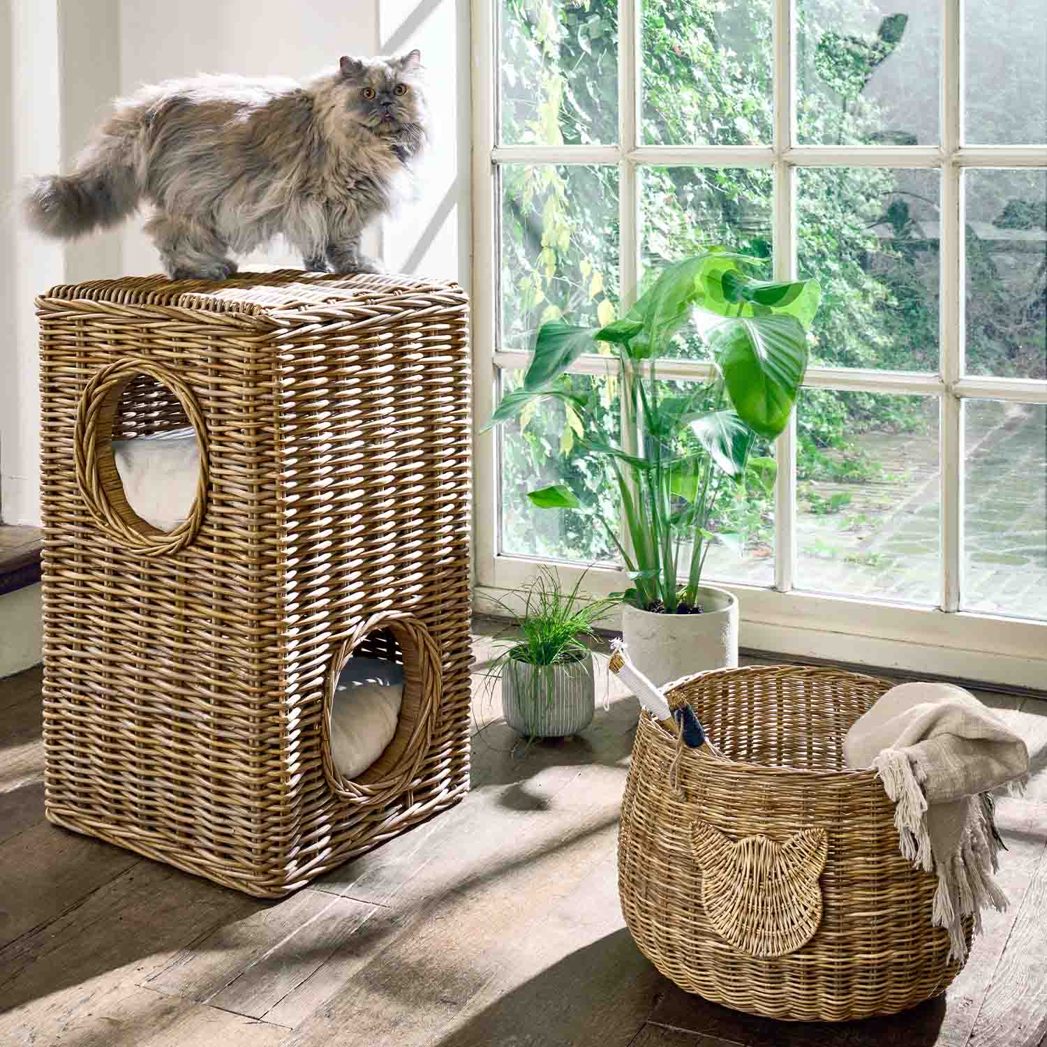 Zweistöckiges Katzenhaus aus natürlichem Rattan mit Kissen neben Wäschekorb und Pflanzen am Fenster