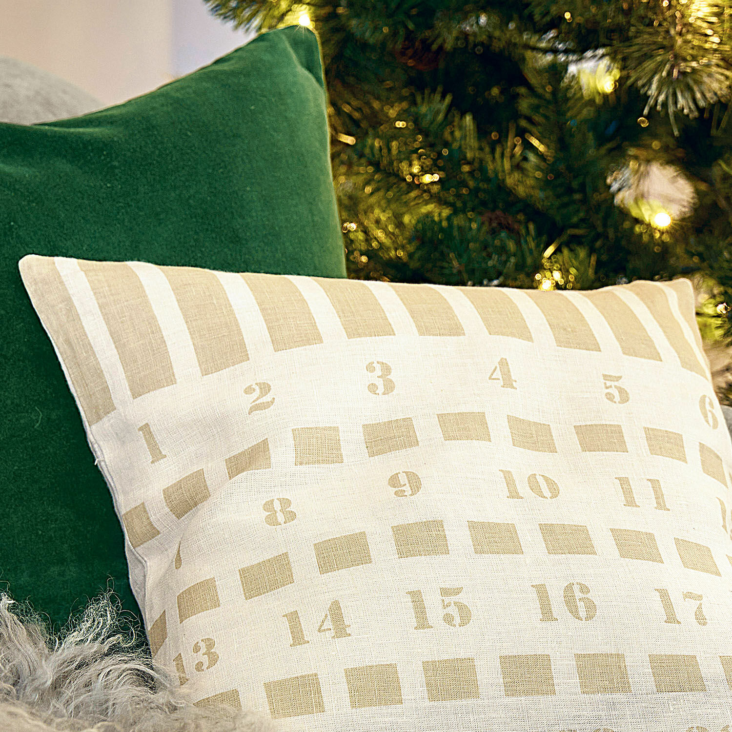 Beige Leinenkissen mit Zahlenmuster vor grünem Kissen und Weihnachtsbaumbeleuchtung