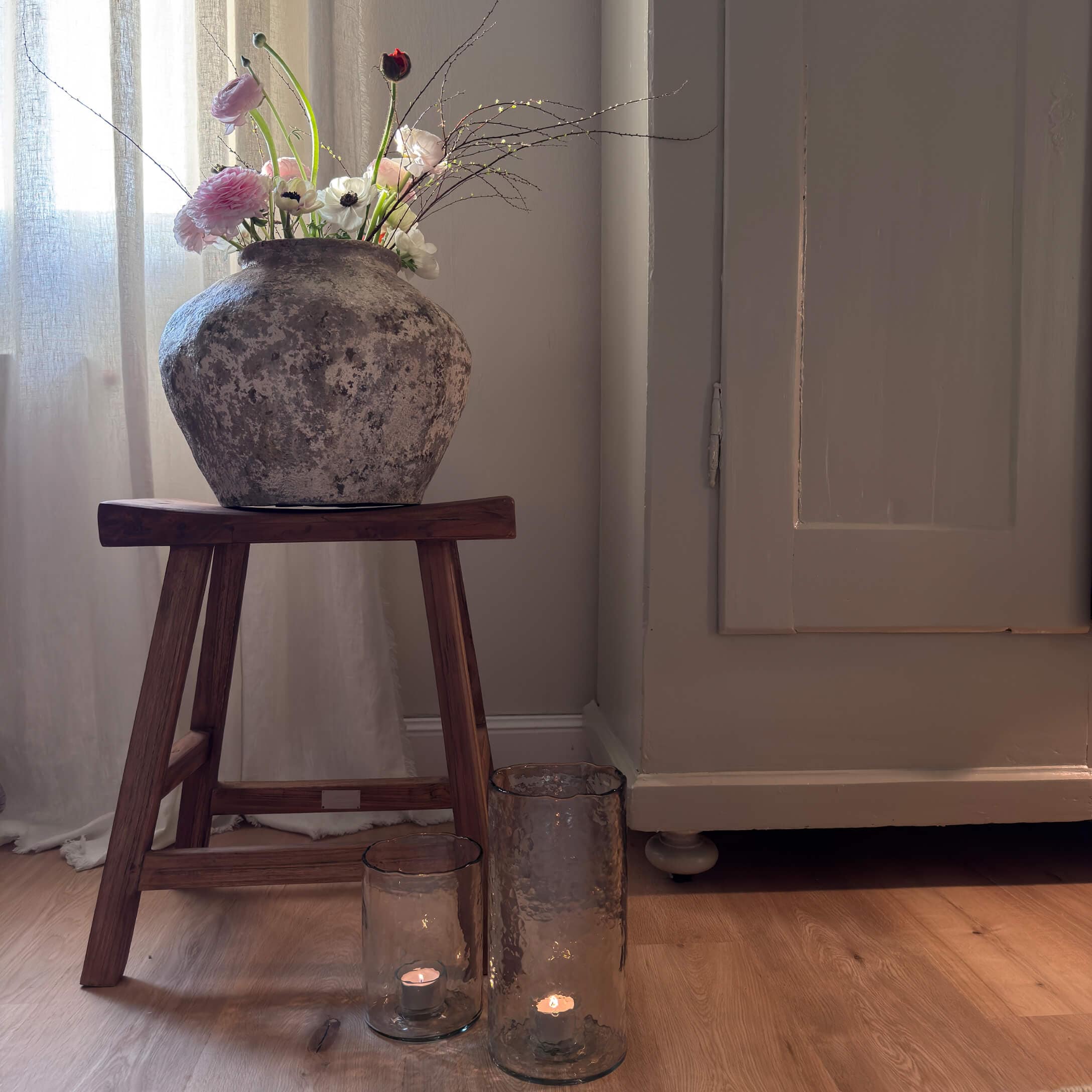 Rustikaler Steinvase mit Blumen auf einem Holzschemel neben Kerzen in Glaslaternen