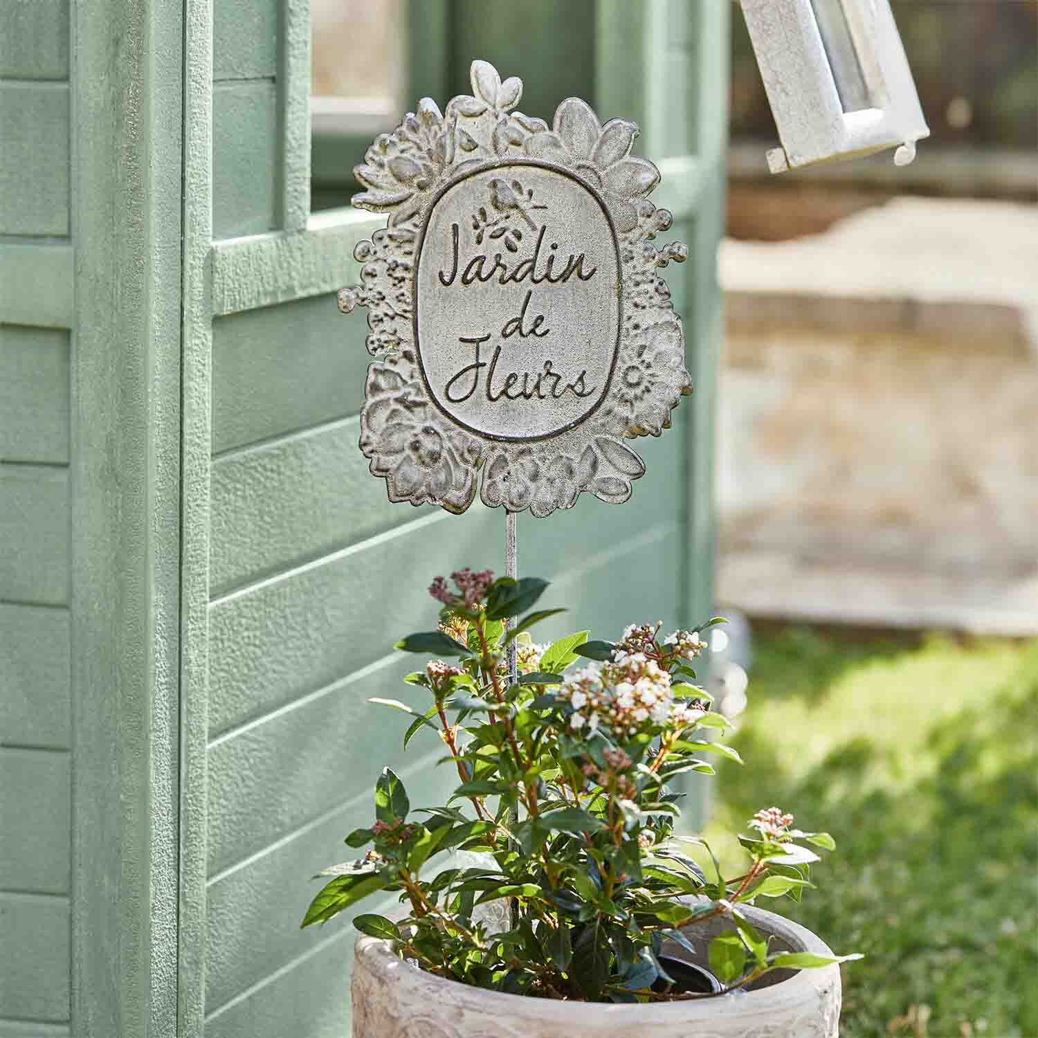Weiße Metall-Gartendekoration mit Schriftzug Jardin de Fleurs vor grünem Hintergrund