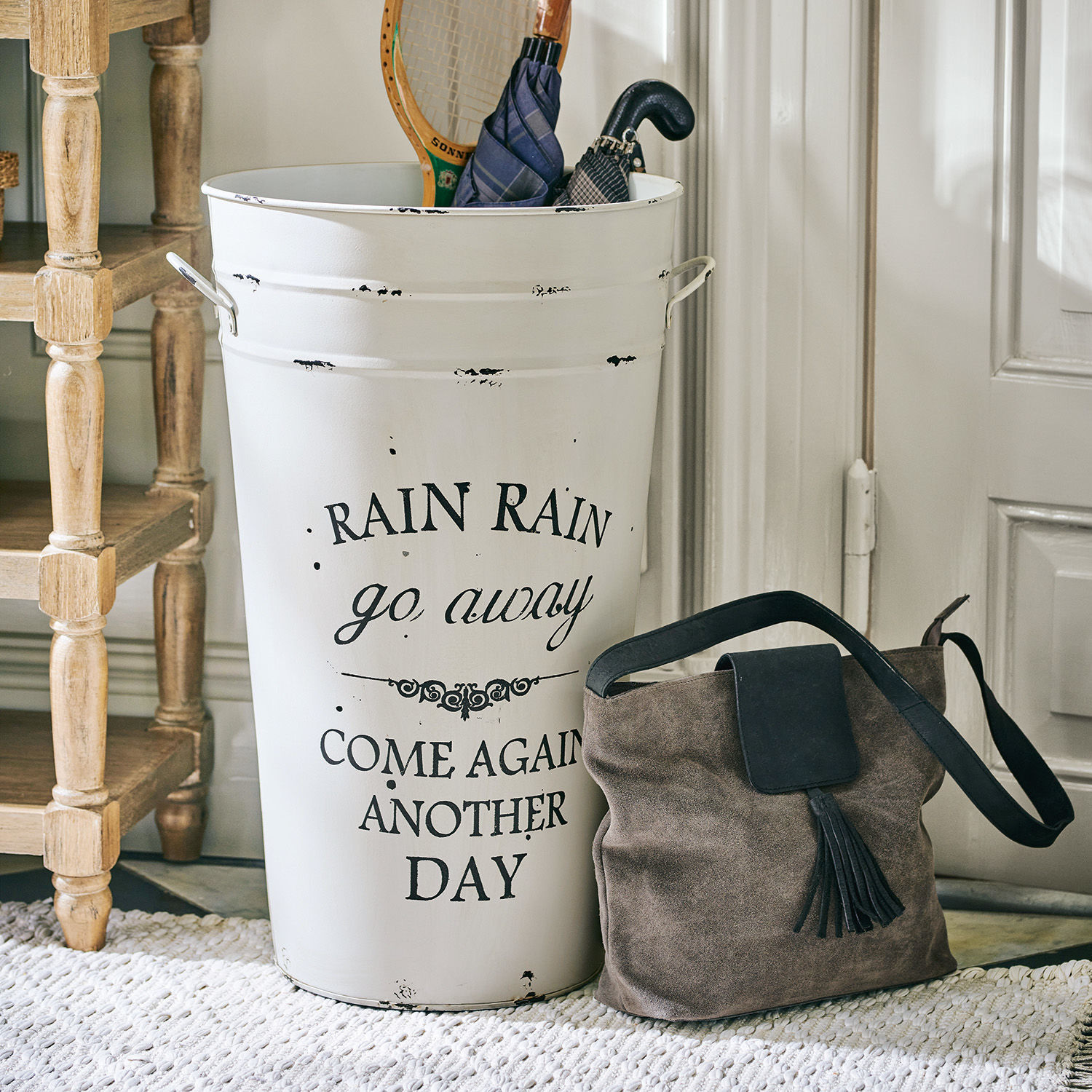 Schirmständer Rain Weißer Metall-Regenschirmständer mit Schriftzug und braune Wildledertasche neben Tür auf Teppich