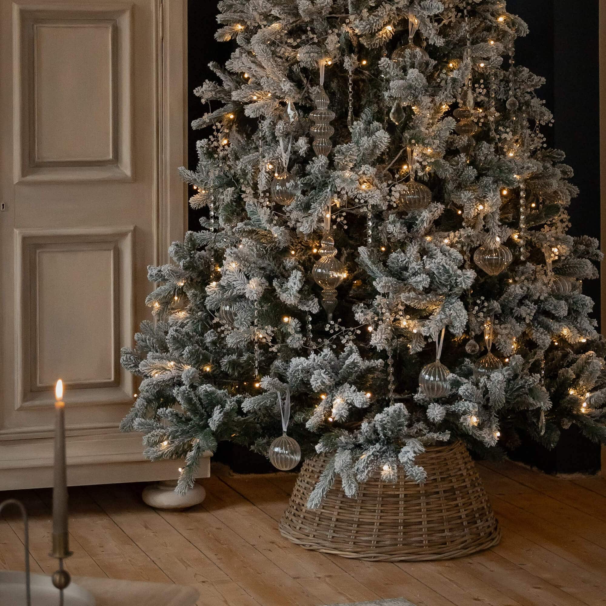 Schneebedeckter Weihnachtsbaum mit Glasornamenten und warmen Lichterketten auf Korbständer