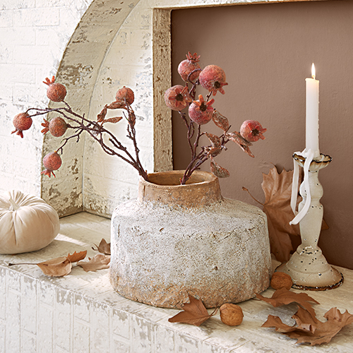 Rustikaler Keramikvase mit Granatästen und weißer Kerze vor herbstlichen Blättern und Kürbis